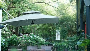 An outdoor seating area with wicker furniture, a large umbrella, and lush greenery. - Blooming Lotus Hostel (Hangzhou)