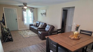 A living room with a brown leather sofa, a wooden dining table with chairs, a ceiling fan, and a window with curtains.