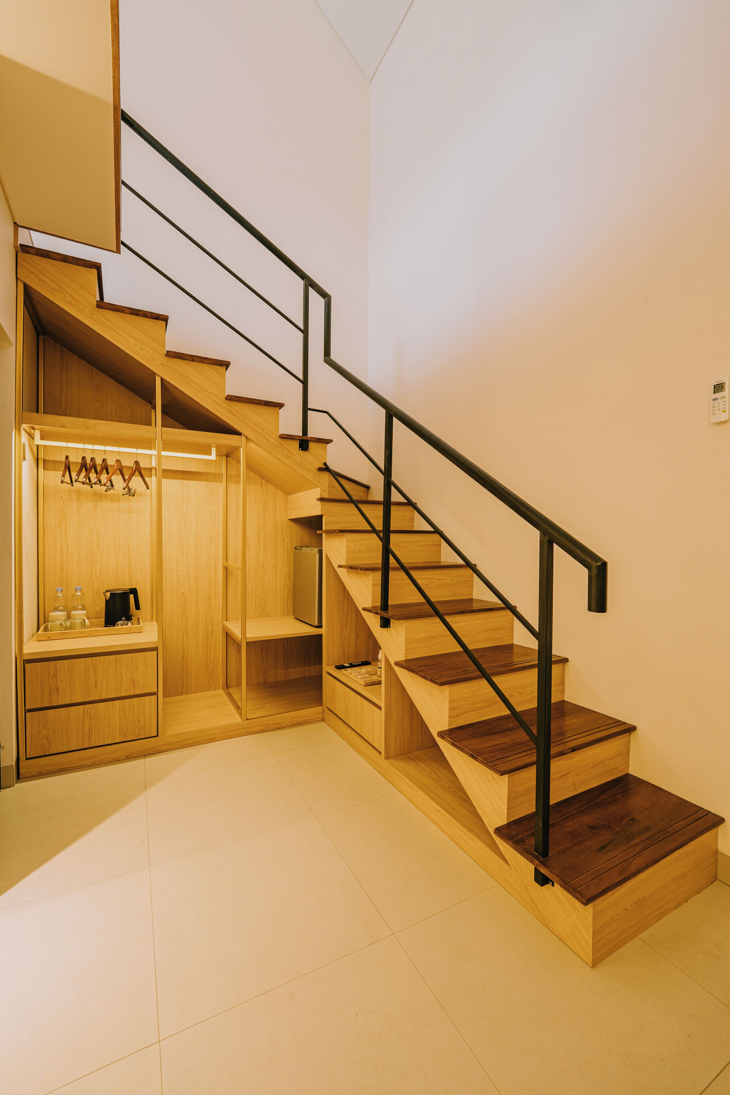Un escalier moderne avec des marches en bois et des rampes noires, menant à un placard en bois doté d’une tringle pour suspendre les vêtements et d’une petite étagère.