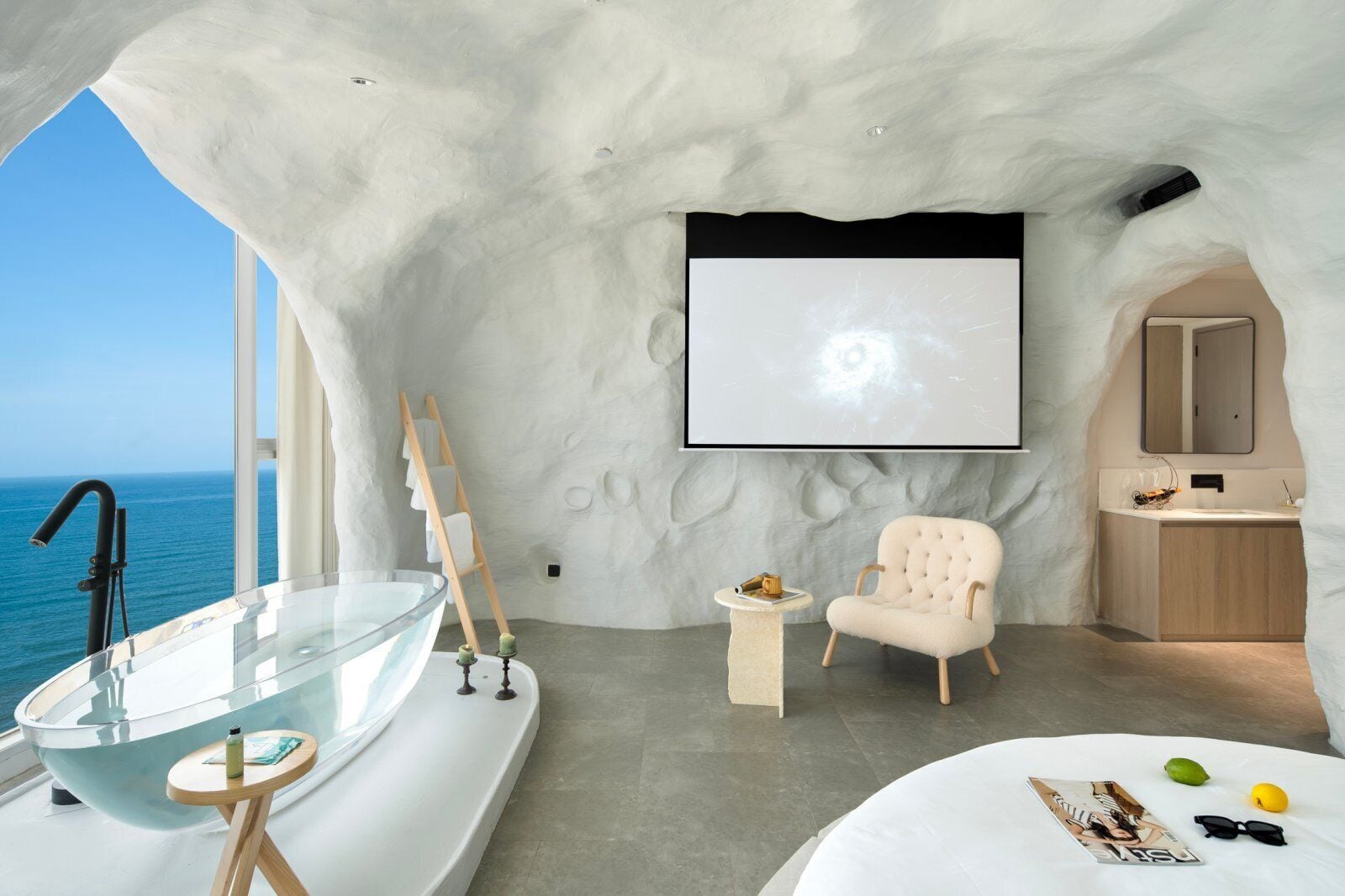 A modern bathroom with a glass bathtub, a white chair, a small table, and a large screen.