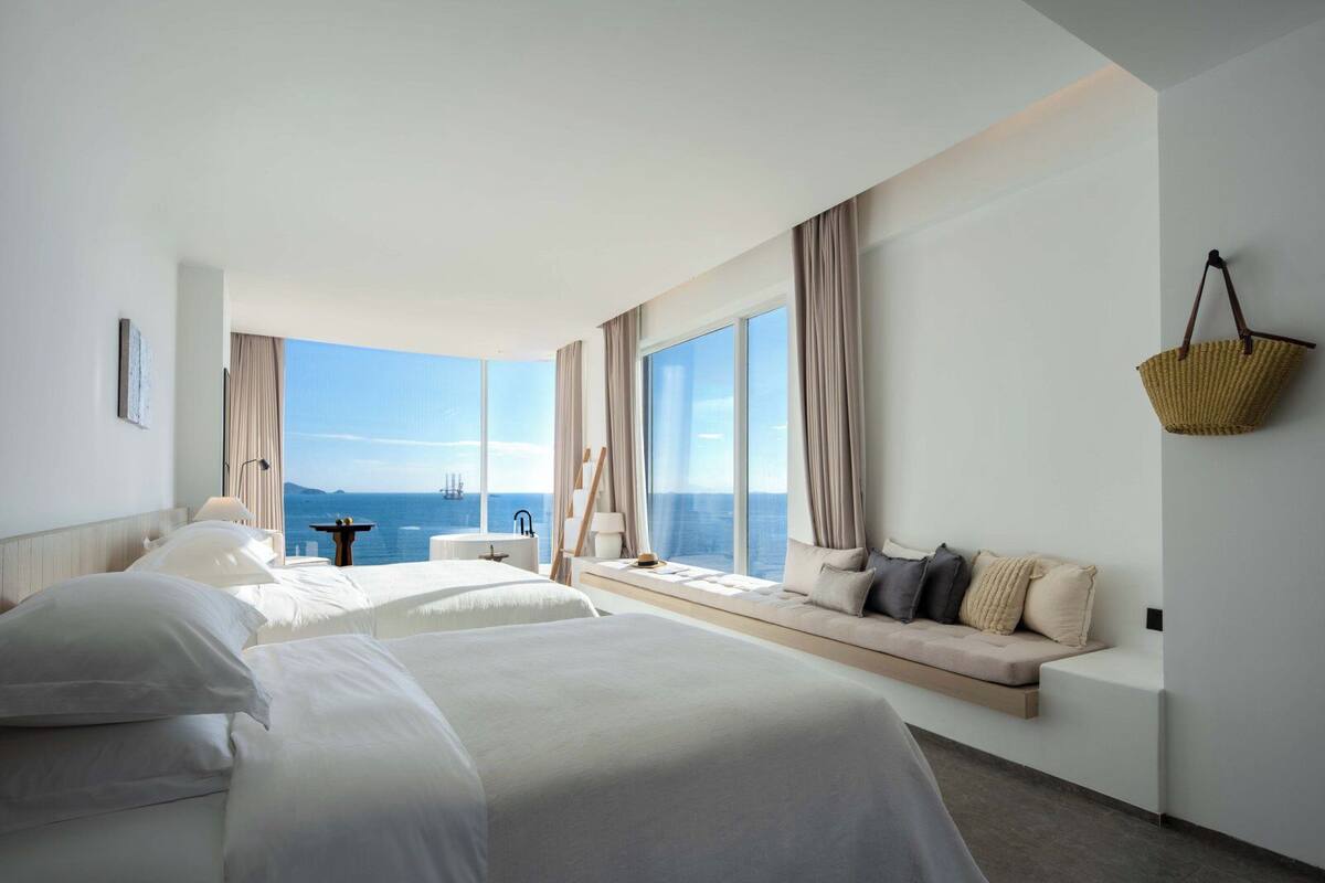 A hotel room with two beds, a large window with a view of the sea, a small table, and a hanging basket.