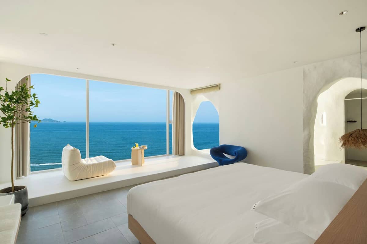 A modern bedroom with a large bed, a seating area by the window, and a view of the sea.