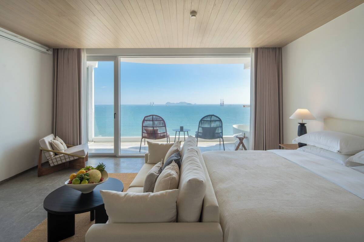 A hotel room with a large bed, a sofa, a dining table with fruit, two chairs, and a view of the sea.