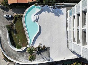 An aerial view of a swimming pool with a curved design, surrounded by a paved area with a few trees and a white building with large windows. - Miyue Santorini Seaview Resort Hotel (Huizhou)