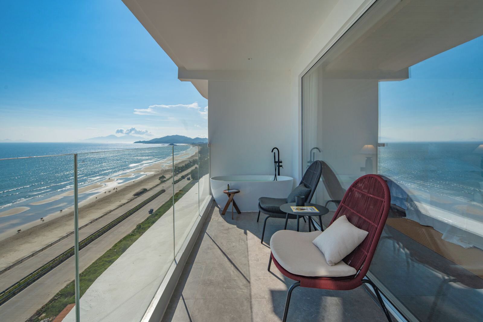 A balcony with a view of the beach and ocean, a bathtub, a chair, and a small table.