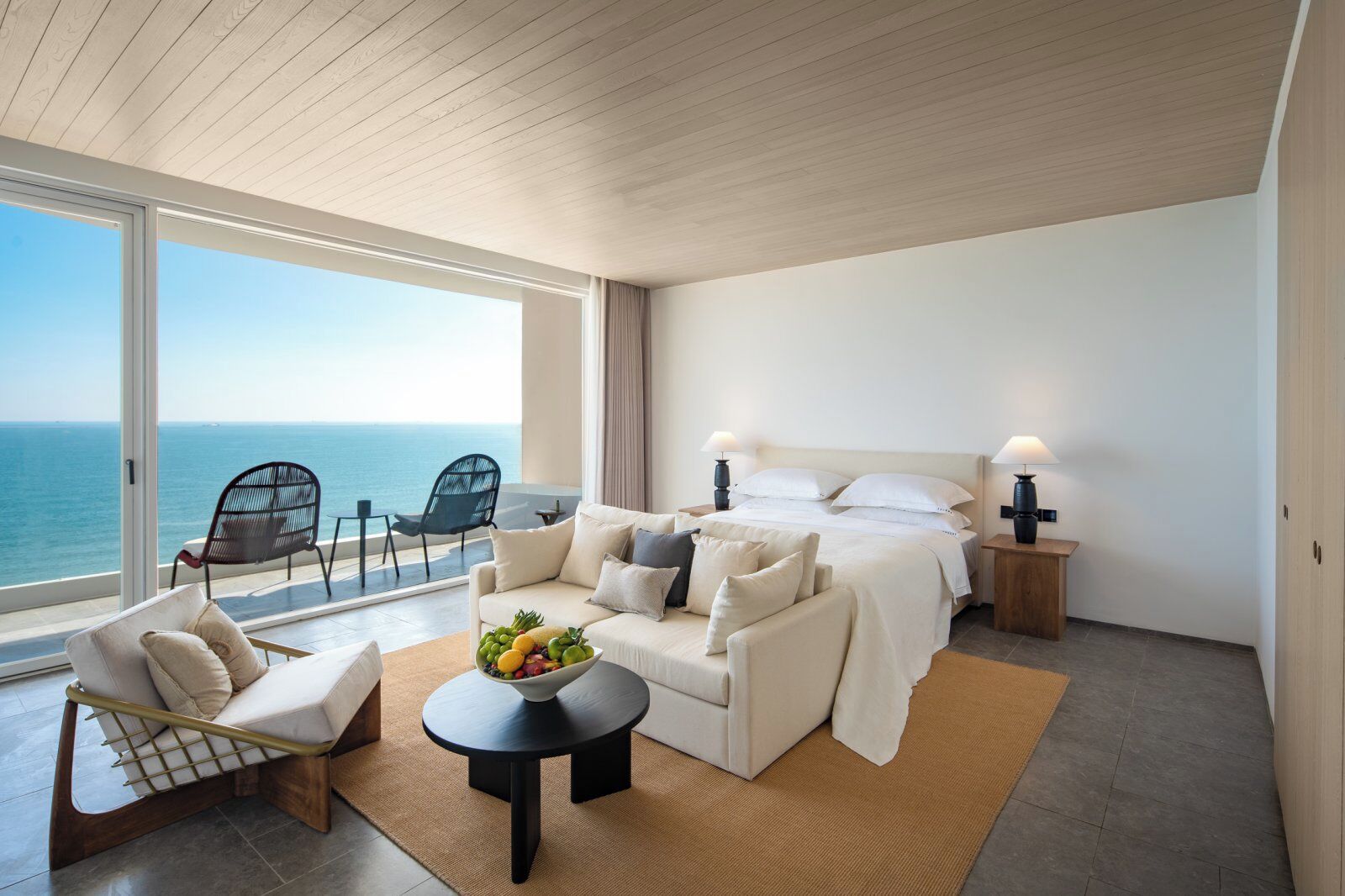 A modern bedroom with a large bed, a sofa, and a dining table. There is a view of the ocean through large windows.