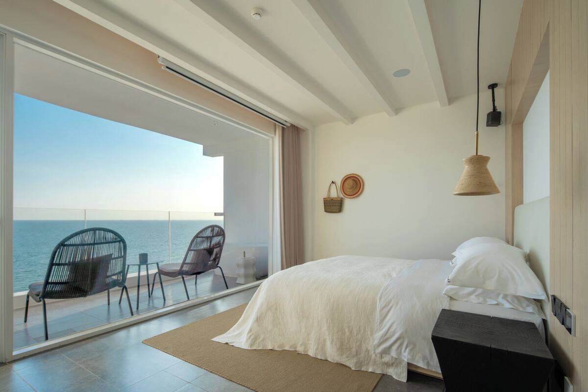 A bedroom with a large bed, a nightstand, a lamp, and a view of the sea through a sliding glass door.
