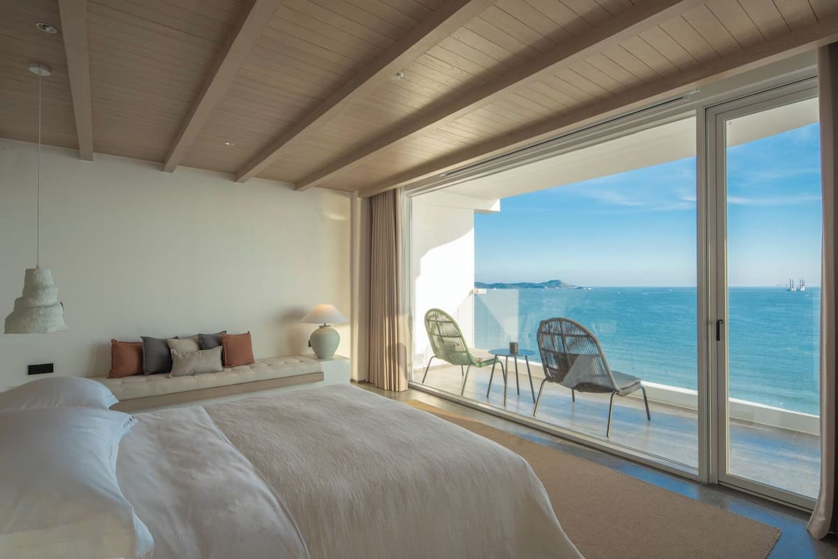 A bedroom with a large bed, a nightstand, a lamp, and a view of the sea through a sliding glass door.