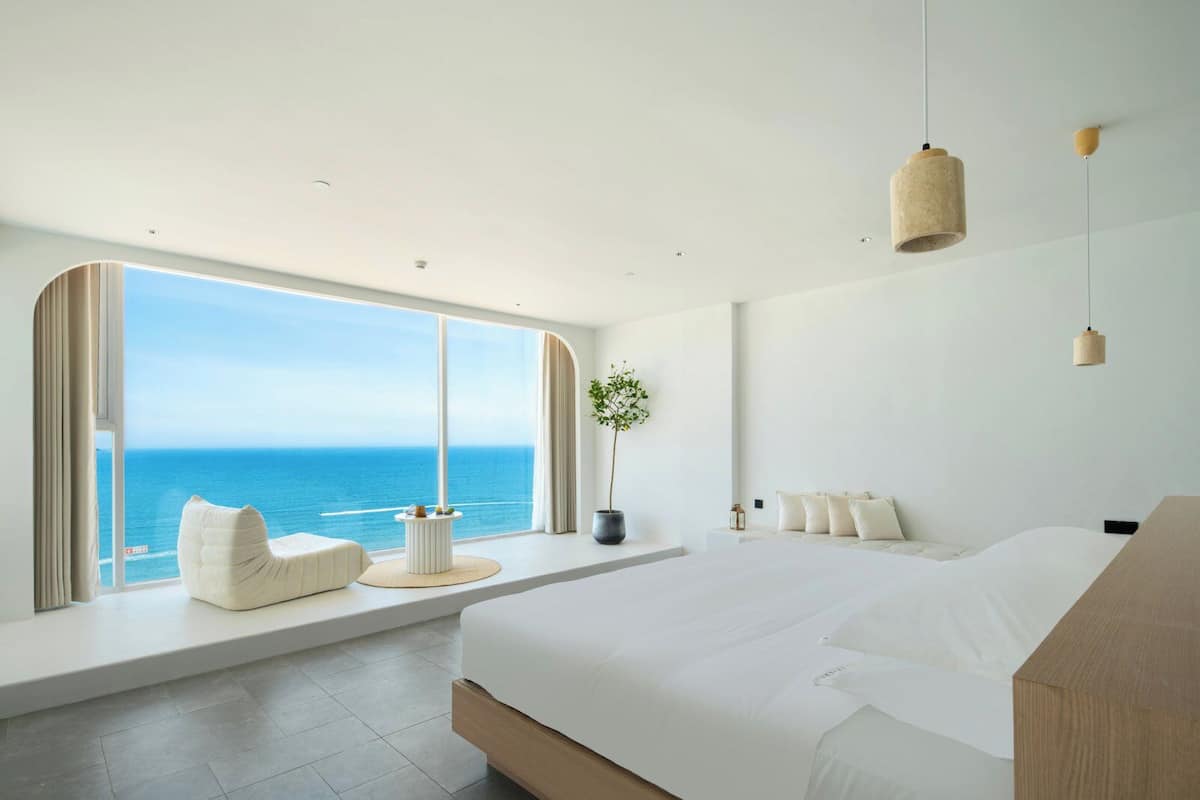 A modern bedroom with a large window overlooking the sea, a bed, a bedside table, and a potted plant.