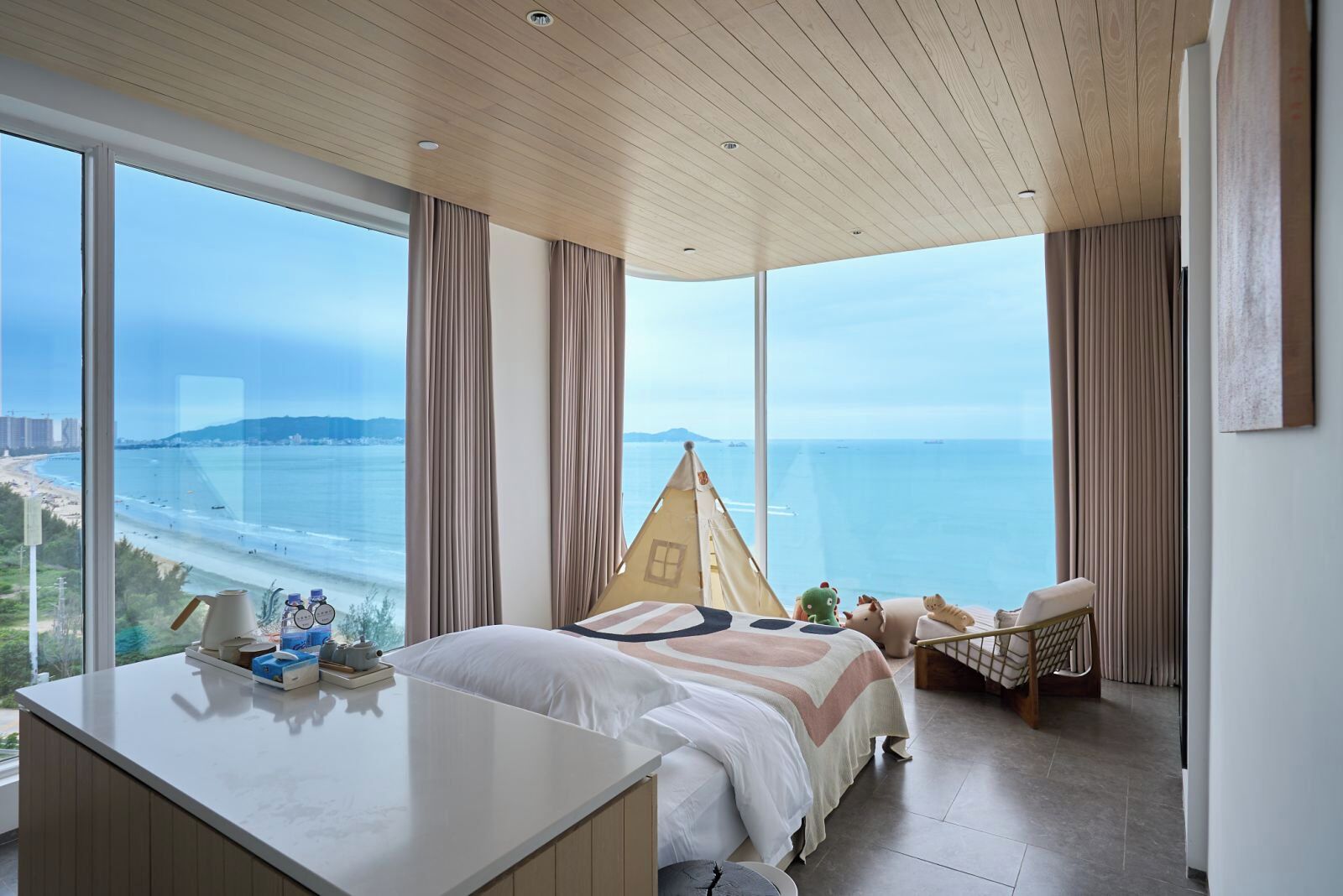 A modern bedroom with a large window overlooking a beach, a bed, a small desk, and a chair.