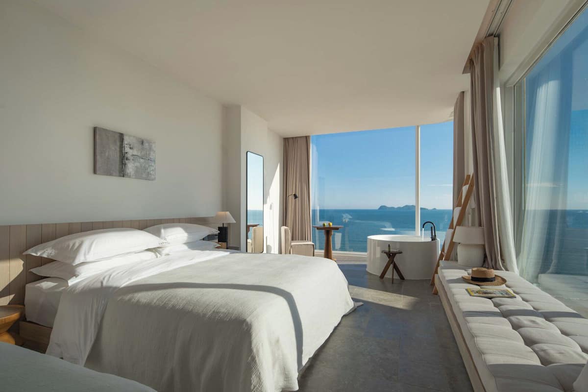 A modern hotel room with a large bed, a sitting area with a view of the sea, and a bathtub.