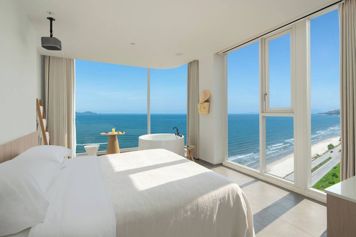 A hotel room with a large bed, a bathtub, a small table with lemons, and a view of the ocean and beach.