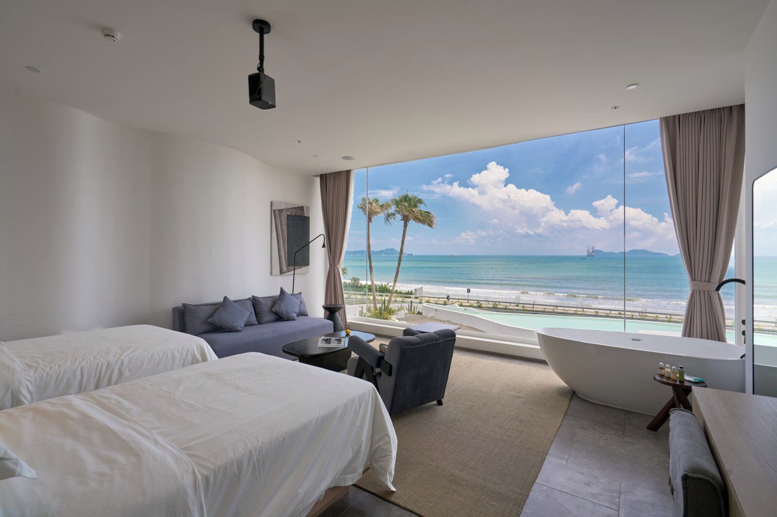 Een hotelkamer met twee bedden, een bank, een kleine tafel, een groot raam met uitzicht op het strand en de oceaan, en een badkuip.