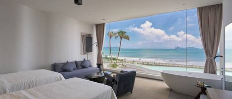 Een hotelkamer met twee bedden, een bank, een kleine tafel, een groot raam met uitzicht op het strand en de oceaan, en een badkuip.