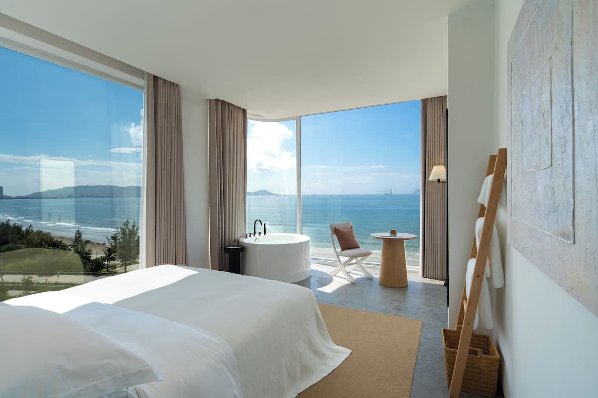 A modern hotel room with a large bed, a bathtub, a chair, and a view of the ocean.