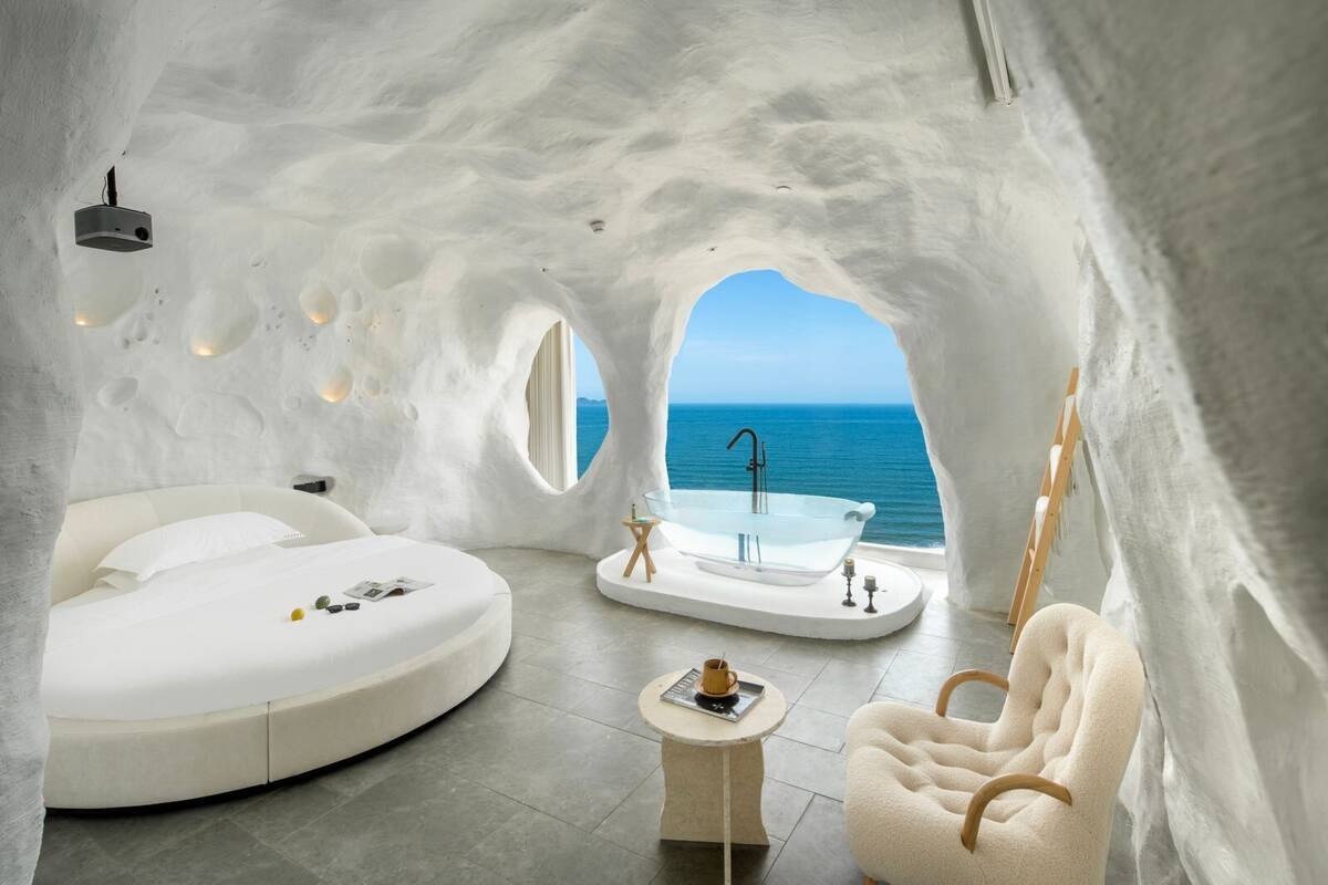 A modern, white-themed interior with a circular bed, a bathtub, and a view of the sea.