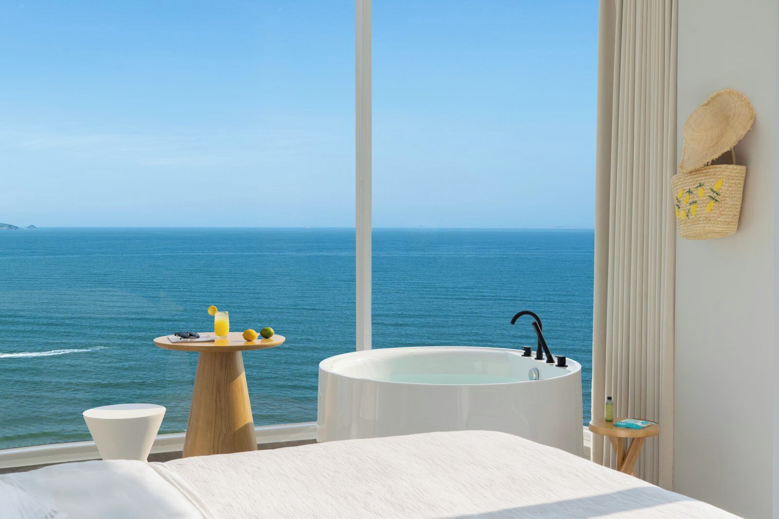 A bedroom with a large window overlooking the sea, a bathtub, a small table with drinks, and a bed.