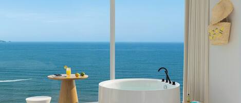 A bedroom with a large window overlooking the sea, a bathtub, a small table with drinks, and a bed.