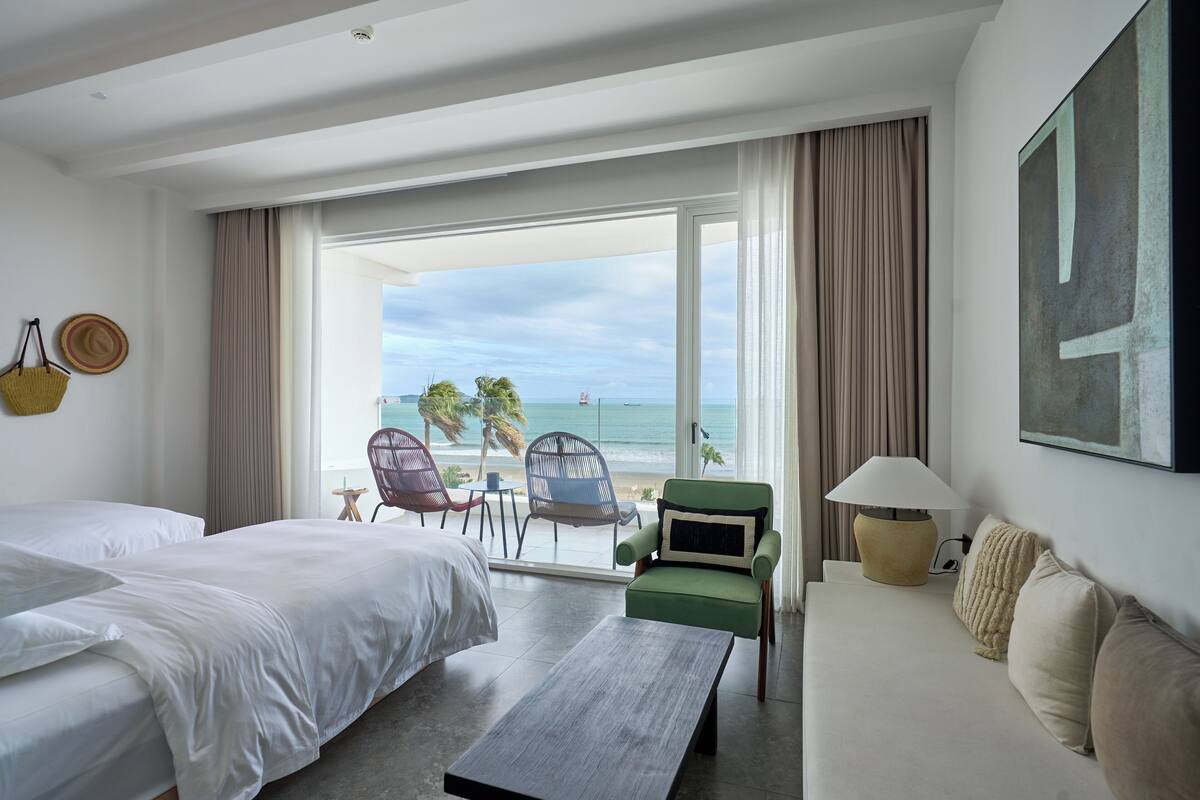 A hotel room with two beds, a green armchair, a sofa, a table, and a view of the beach.