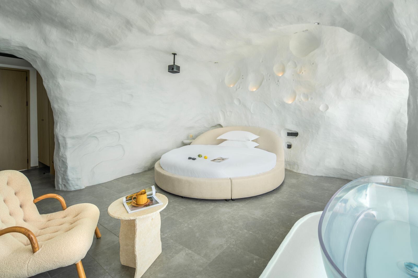 Un dormitorio moderno con una pared original de estilo hielo, una cama redonda, una bañera de vidrio y un sillón acogedor.