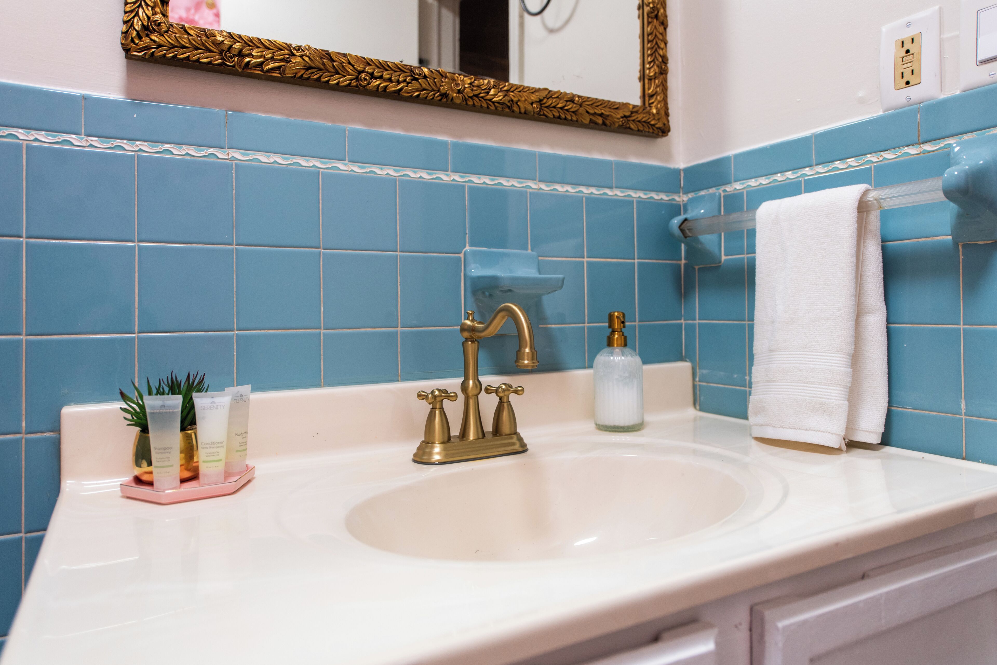 Un baño con lavamanos blanco, azulejos azules, un espejo con marco dorado y una toalla blanca colgada en un perchero.