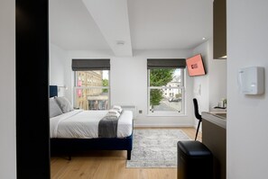 Une chambre d’hôtel moderne équipée d’un lit, d’un bureau, d’une chaise, d’une fenêtre et d’un téléviseur fixé au mur.