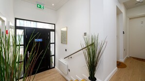 Un couloir avec une porte noire, un panneau de signalisation verte indiquant la sortie, un tableau encadré et une plante en pot.