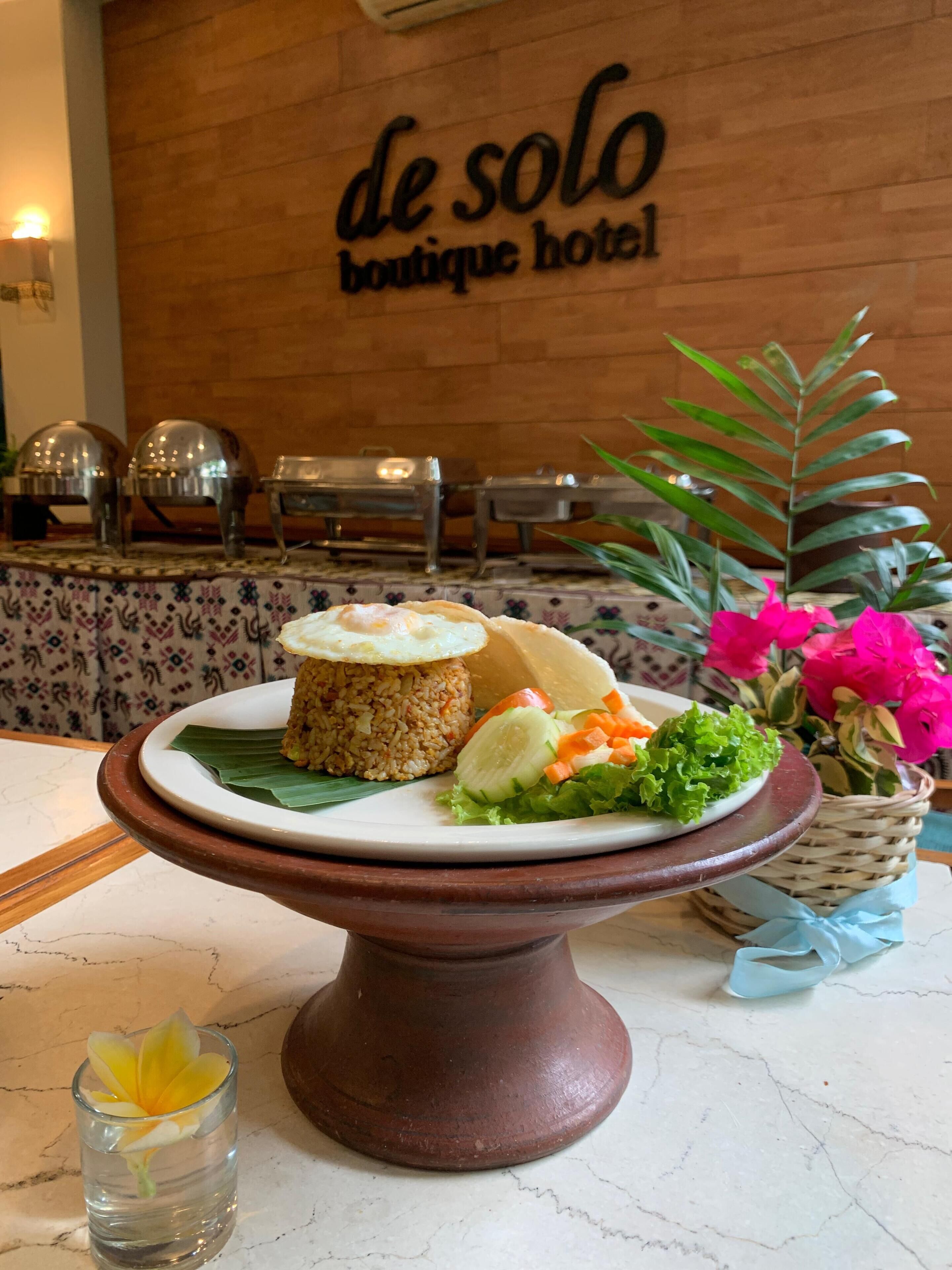Une table à manger avec une assiette de nourriture, un petit verre avec une fleur, et, en arrière-plan, une enseigne indiquant « de solo boutique hotel ».