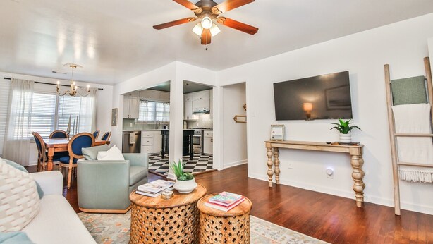 A spacious living room with a ceiling fan, a dining area with blue chairs, a kitchen with white cabinets, and a TV mounted on the wall. - Cute Bungalow in Central Lubbock (Lubbock)