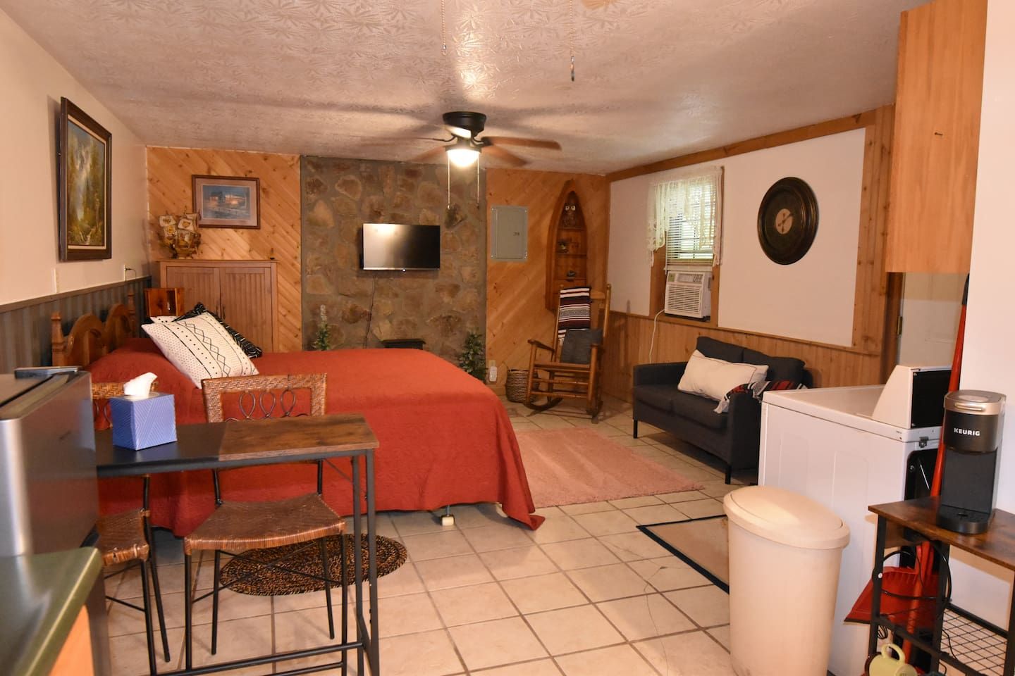 A compact room with a bed, a small table, a chair, a sofa, a television, a ceiling fan, and a washing machine.