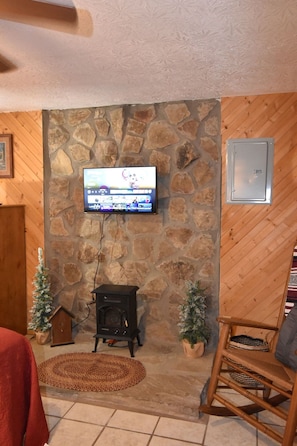 Un mur en pierre avec un téléviseur fixé au-dessus d’une cheminée, une chaise en bois et un petit sapin de Noël.