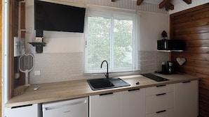 A compact kitchen with a built-in dishwasher, a sink, and a window with blinds. - L'Échappée Créole (Baie-Mahault)