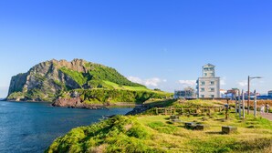 A coastal landscape with a prominent rocky island and a modern building with a tower. - Sunrise Castle Pension (Seogwipo)