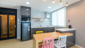 A compact kitchen with a refrigerator, microwave, and sink. There is a dining table with chairs and a window with blinds. - Sunrise Castle Pension (Seogwipo)