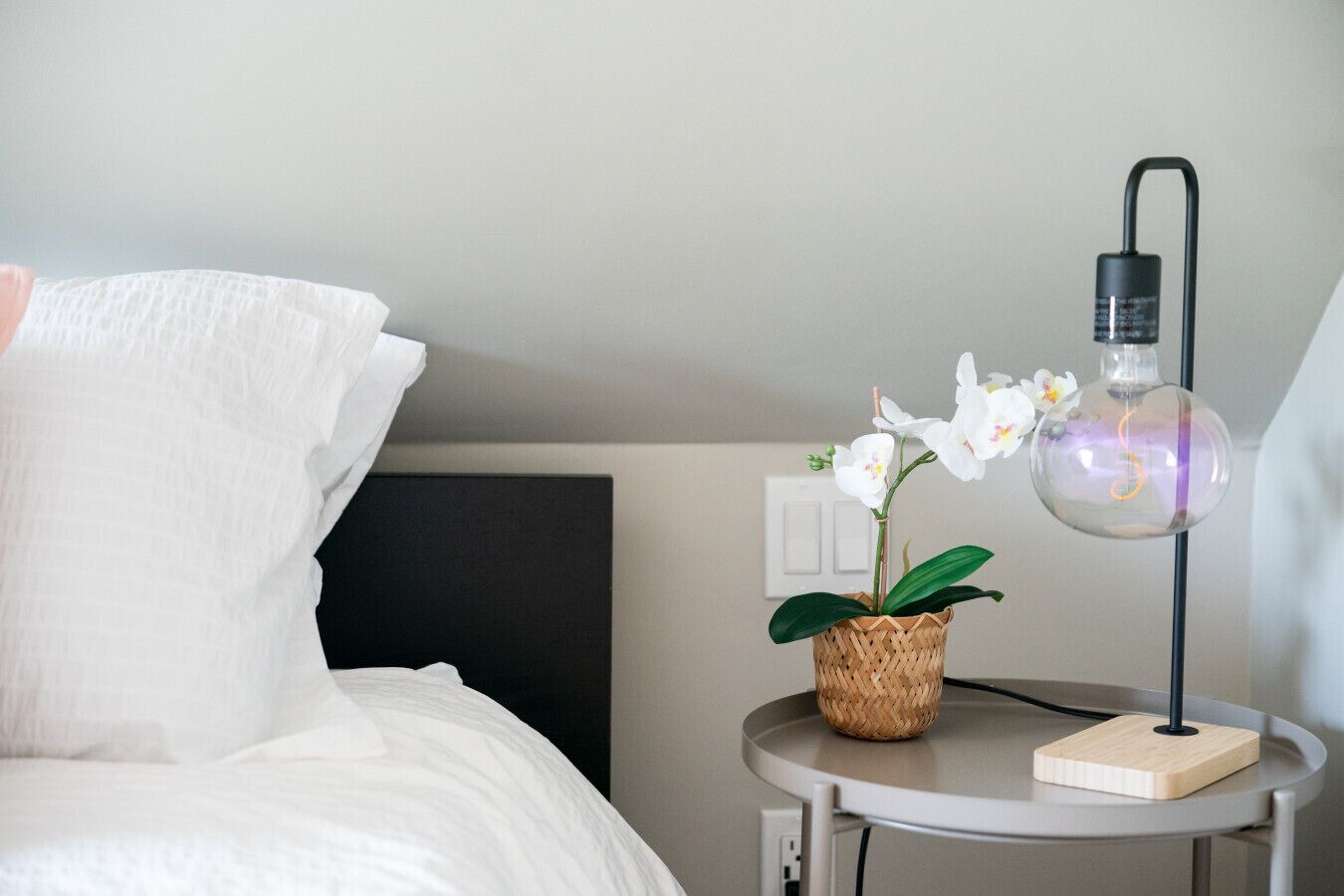 Un lit bien fait, avec une literie blanche, une petite table ronde sur laquelle se trouve une lampe et un pot d’orchidée, et une tête de lit sombre.