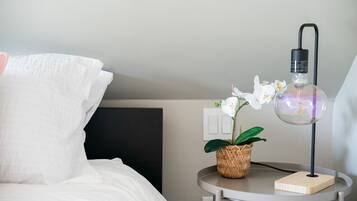 Un lit bien fait, avec une literie blanche, une petite table ronde sur laquelle se trouve une lampe et un pot d’orchidée, et une tête de lit sombre.