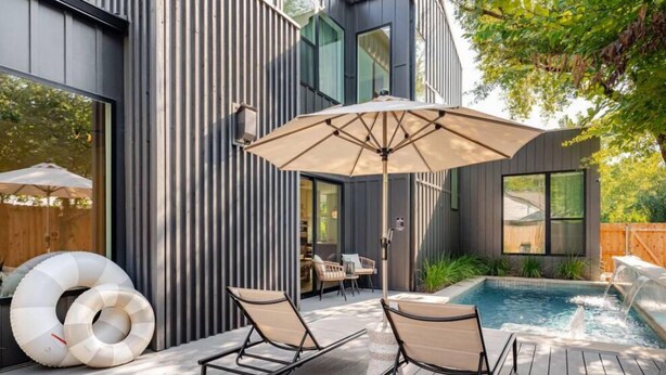 A modern outdoor deck with two lounge chairs and a large umbrella, a swimming pool with a waterfall feature, and a wooden fence in the background.