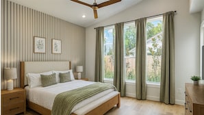 A bedroom with a large bed, two bedside tables, a ceiling fan, and a view of the outdoors through a window with curtains.