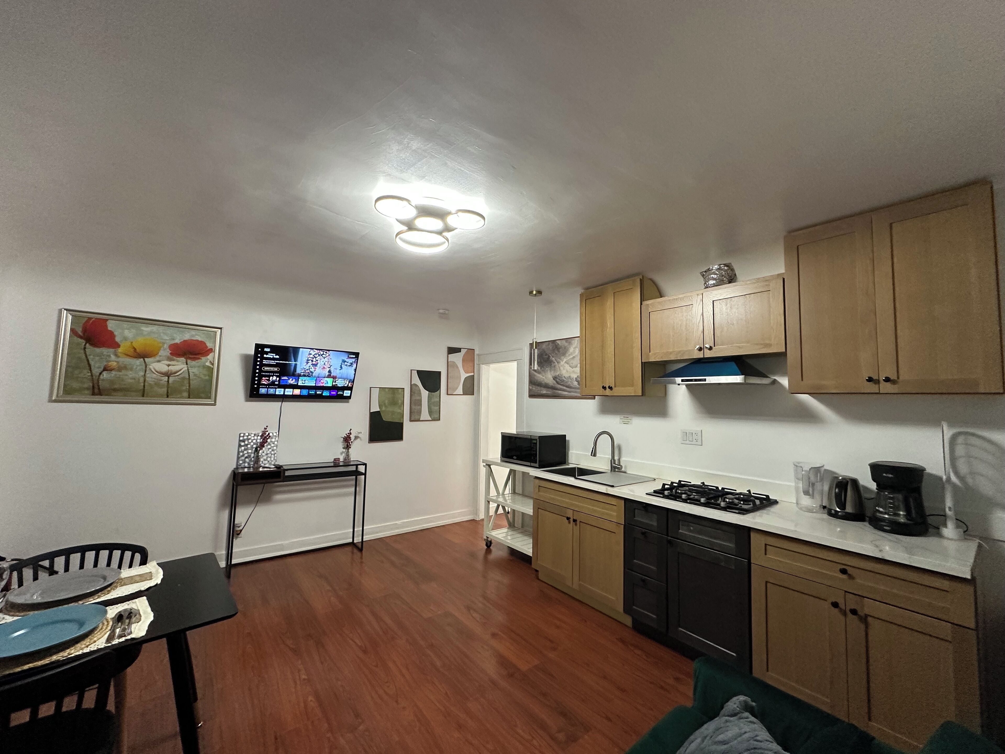 A compact kitchen with wooden cabinets, a microwave, a sink, and a stove. There is a dining table with chairs and a TV mounted on the wall.