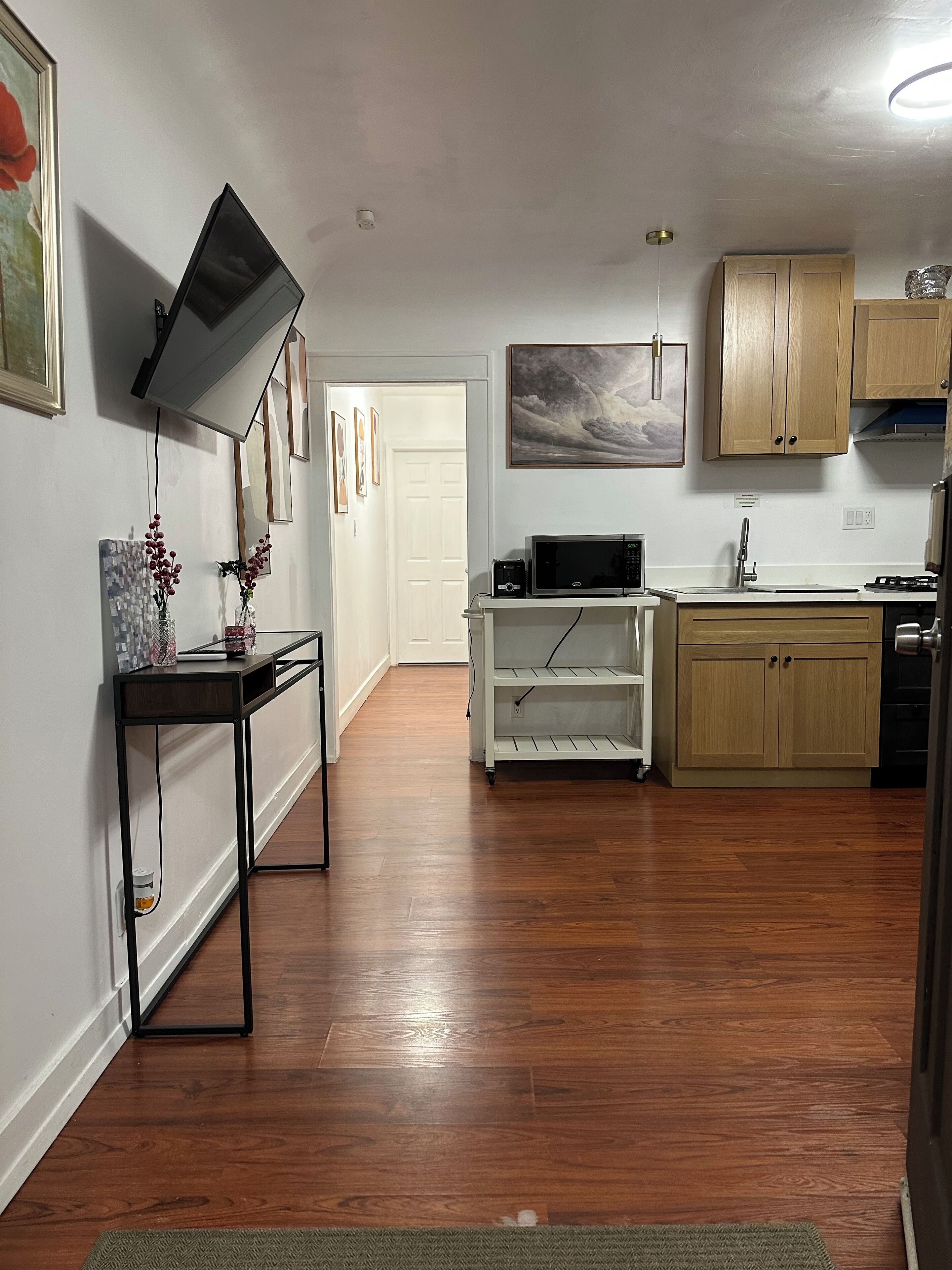 A modern kitchen with wooden cabinets, a microwave, and a sink. There is a TV mounted on the wall and a small table with decorative items.