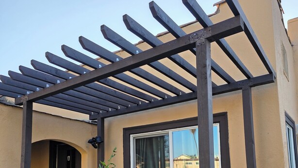 Un espacio de estar al aire libre cubierto con una pérgola, una mesa de vidrio y una silla de mimbre.