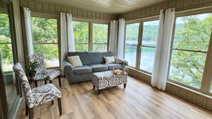 Amplia sala de estar con un sofá gris, sillones estampados y piso de madera. Grandes ventanales ofrecen vistas a un lago y vegetación.
