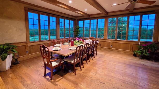 A dining room with a large wooden table set for six, surrounded by wooden chairs. The room has a high ceiling with exposed beams and a ceiling fan. Large windows provide a view of the outdoors.