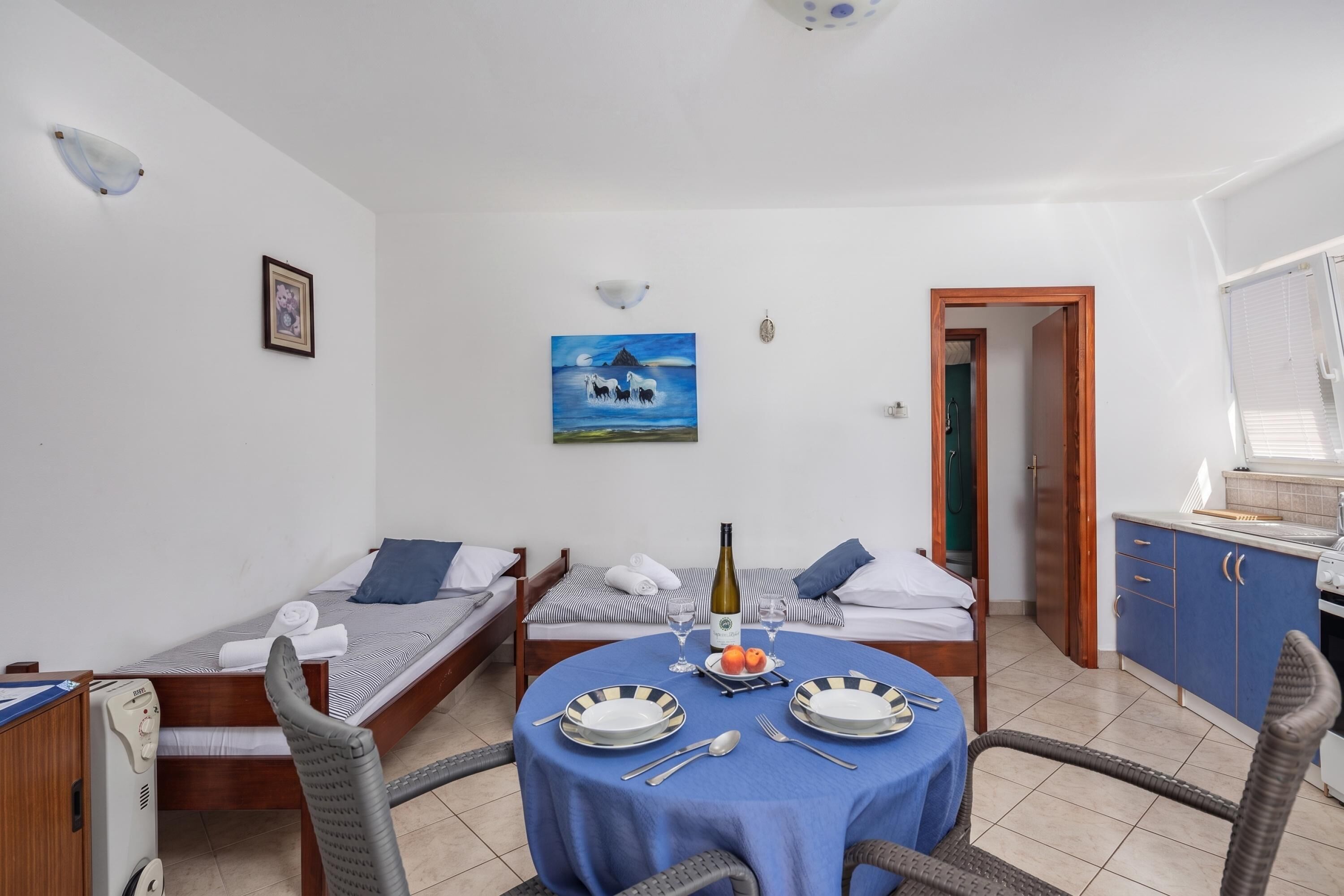 A compact hotel room with two single beds, a dining table with chairs, a small kitchen area, and a painting on the wall.