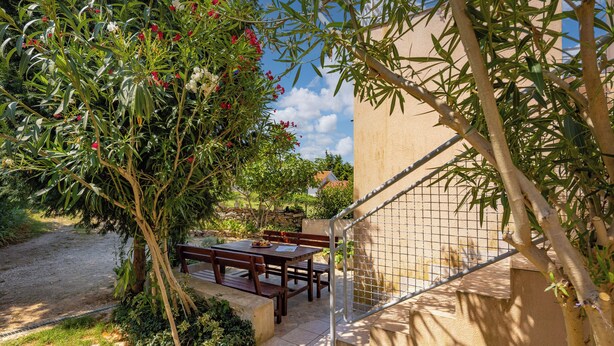 Een terras buiten met een houten tafel en bankjes, omgeven door groen en een gevel.