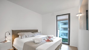 Quarto de hotel moderno com uma cama grande, uma mesa de cabeceira, uma luminária para leitura e vista da cidade por uma janela ampla.
