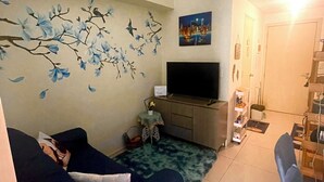 A living room with a sofa, a flat-screen TV, a coffee table, and a wall with a floral mural. - Cosy Condo with Balcony at Makati SM Jazz (Makati City)