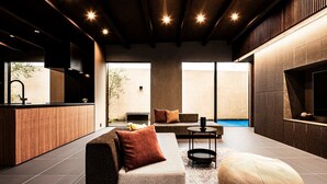 A modern living room with a sofa, ottoman, and a small coffee table. There is a kitchen area with a sink and a window overlooking a pool. - A private villa where you can step away from your daily life and savor "extra... (karatushi watada honmura)