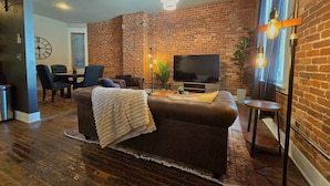 A living room with a brick wall, a sofa, a TV, and a dining area with chairs.