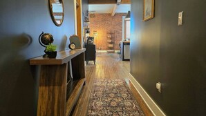 A hallway with a wooden console table, a mirror, and a patterned rug leading to a room with a brick wall and a desk.